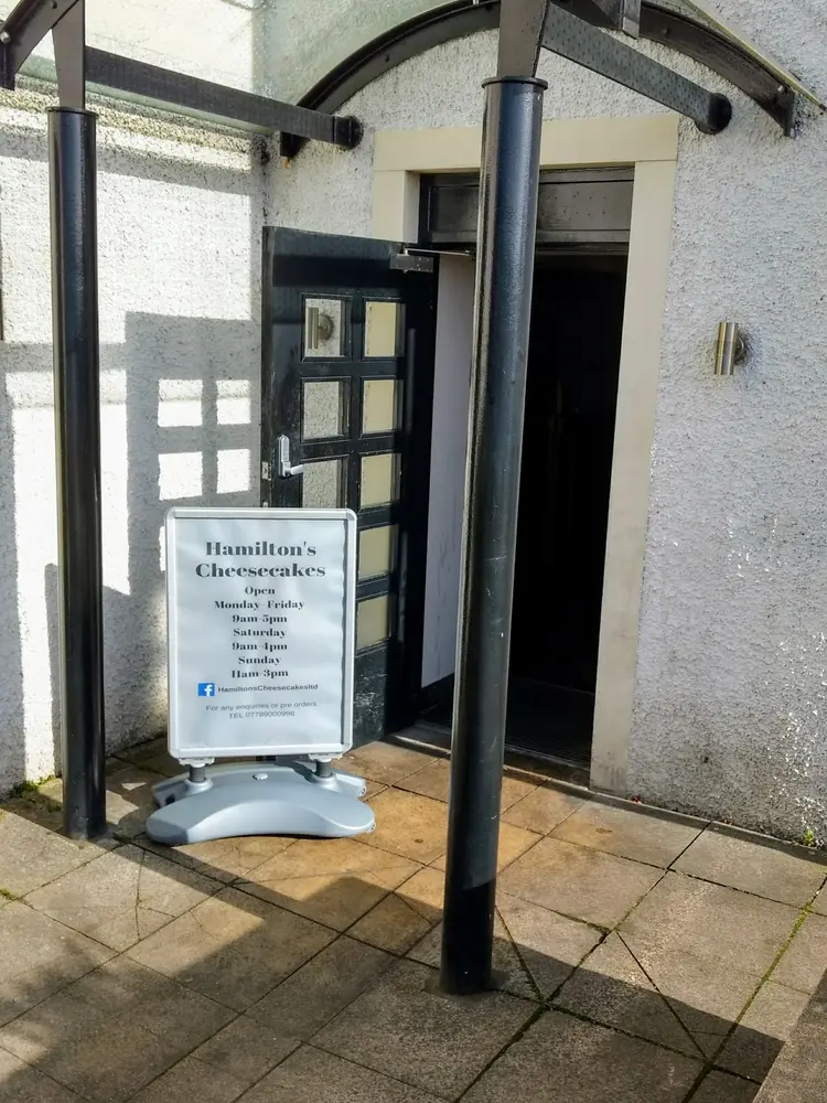 Shop doorway under a glass canopy with a Hamilton's Cheesecakes opening-hours board outside